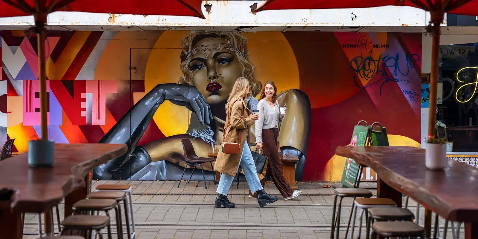 Two women strolling through Christchurch in front of a mural, holding coffee