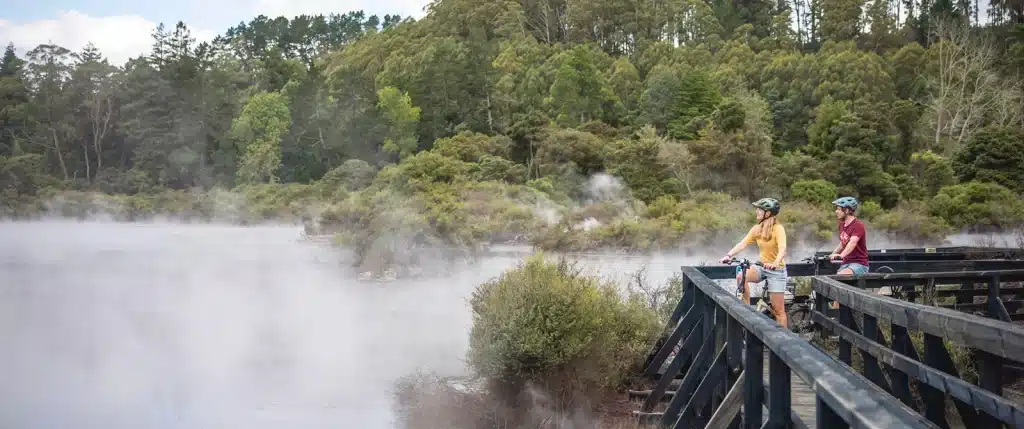 Couple bicycling through a geothermal park in New Zealand, with steam rising out of the lake