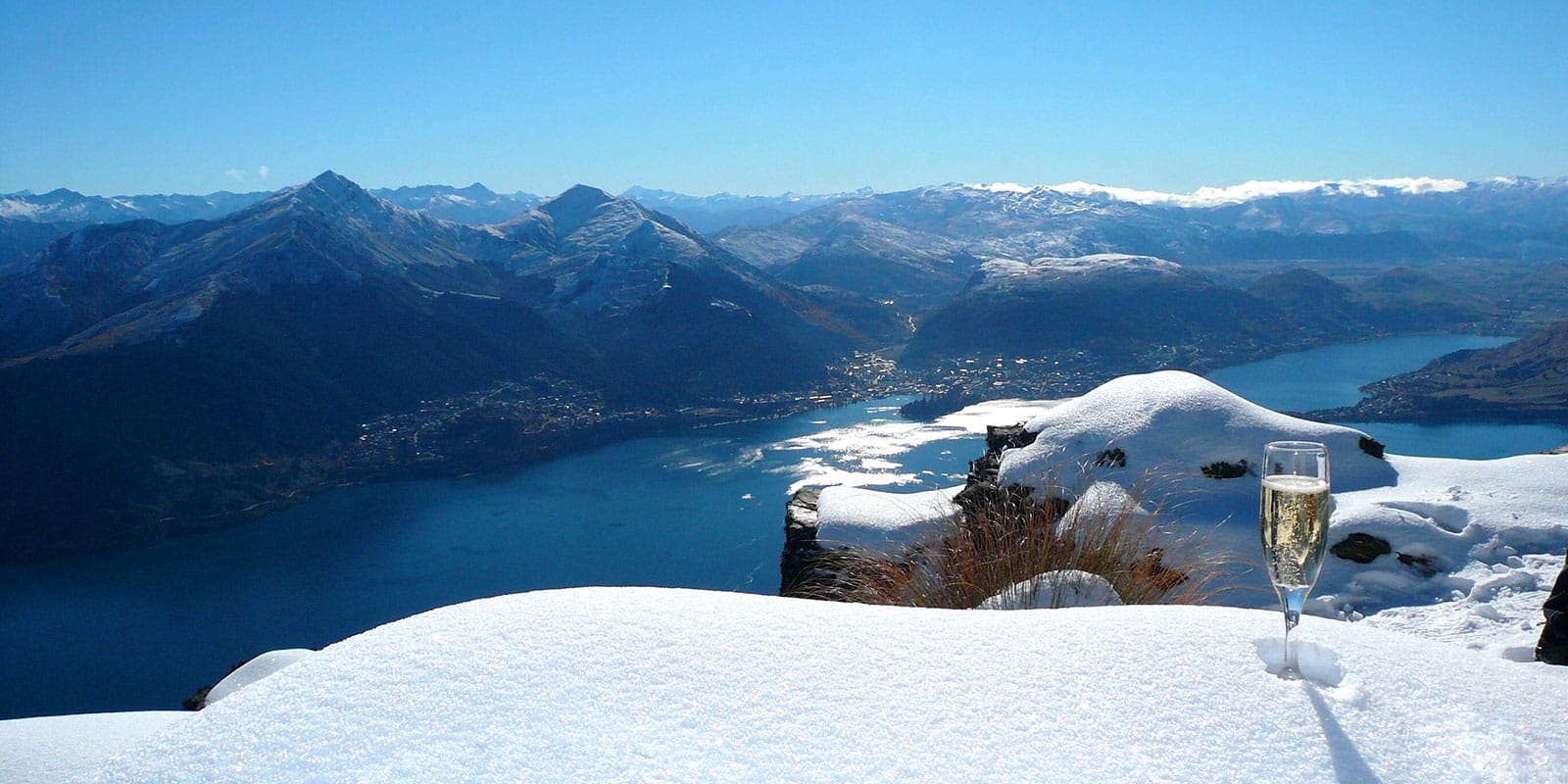 Glass of champagne on the snow, with a panoramic view of Lake Wakatipu and surrounding mountains